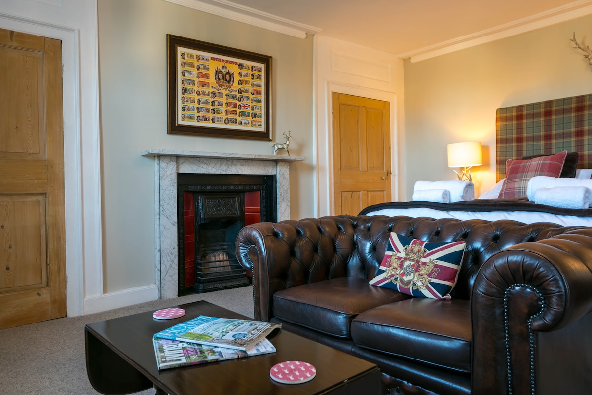 A spacious double room features a large, comfortable king-sized bed and a traditional chesterfield sofa adorned with a Union Jack cushion. A vintage marble fireplace adds character, while a piece of framed wall art enhances the decor. A wooden door leads to another area.
