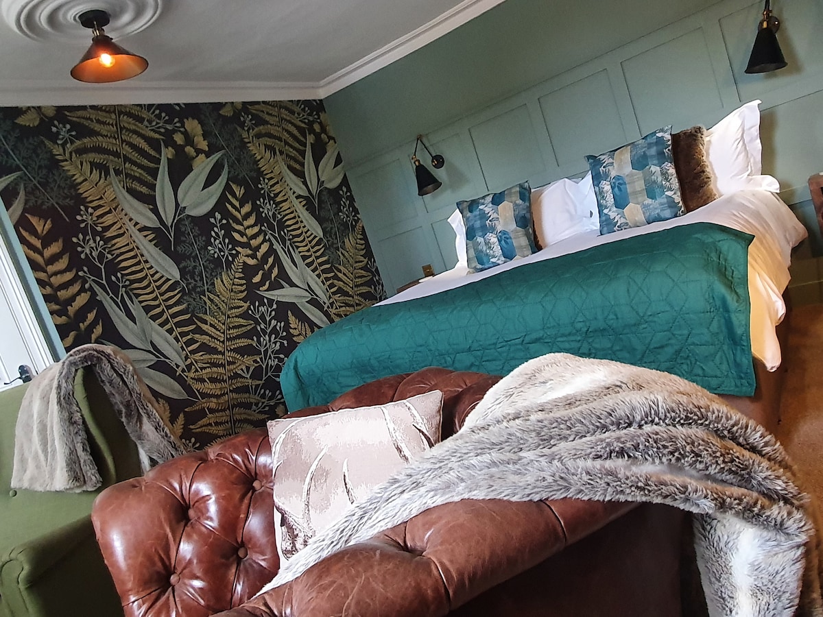 A spacious bedroom features a sumptuous bed layered with a teal throw and decorative cushions. The accent wall is adorned with a botanical pattern, while a leather sofa adds a cozy touch. Soft lighting from wall sconces complements the rustic decor.