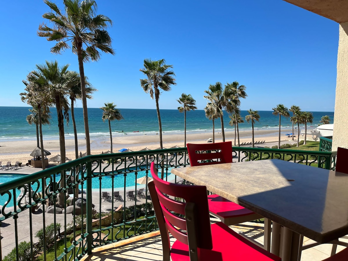 A private balcony is featured, showcasing a clear view of the sandy beach and ocean. Bright red chairs surround a stone table, while palm trees line the shore, creating a natural frame for the tranquil scene. Sun and blue skies complete the coastal atmosphere.
