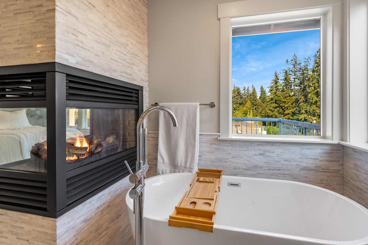 A modern bathroom features a freestanding bathtub positioned beside a stylish fireplace. Large windows provide a serene view of the surrounding trees, while a wooden bath tray rests across the tub, ready for relaxation.
