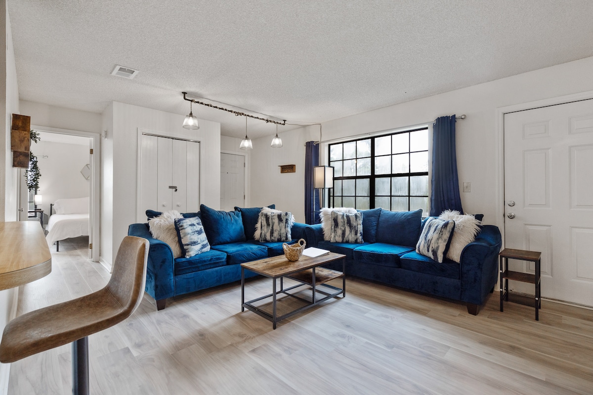 A spacious living area showcases a large blue sectional sofa adorned with decorative pillows. A coffee table sits in front, and natural light filters through the sizable window, complemented by light curtains. A subtle bar stool is positioned nearby, enhancing the open design.