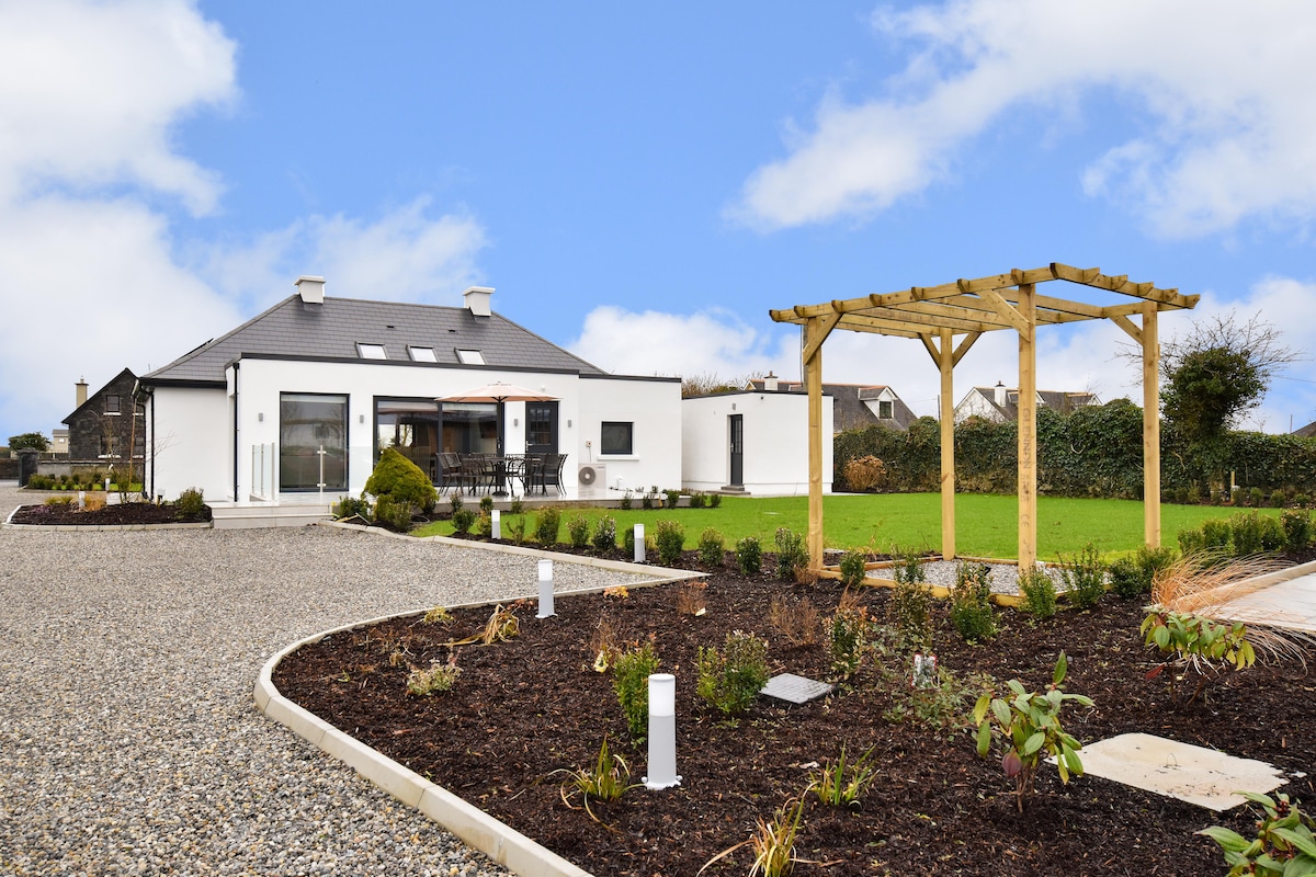 The newly renovated bungalow is surrounded by landscaped gardens, featuring a timber pergola. A spacious patio area is visible, with sliding glass doors leading into the interior. A gravel driveway provides access to the house, enhancing the serene country setting.