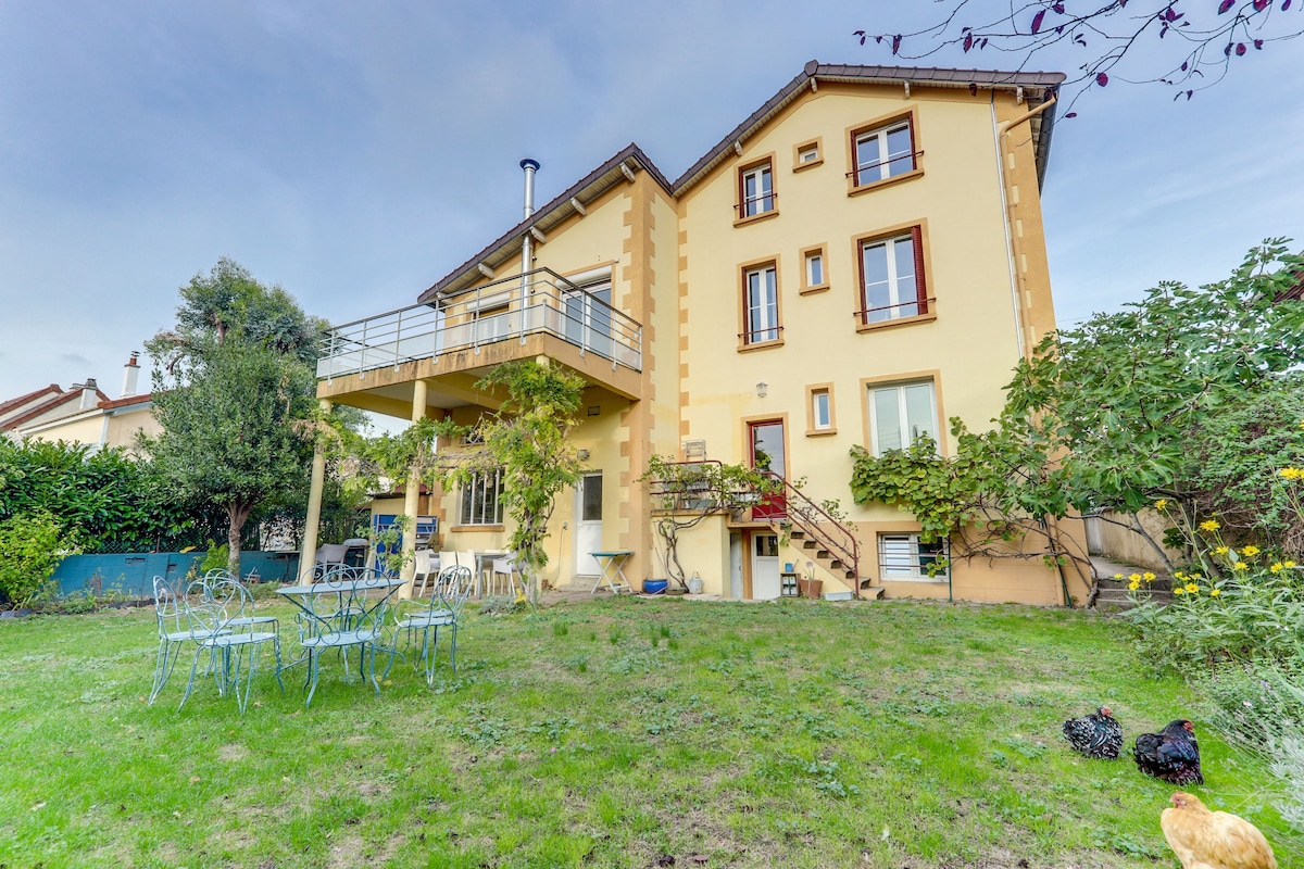 The house presents a charming exterior with multiple stories and a terrace overlooking the garden. A well-maintained lawn features several outdoor chairs arranged around a table, surrounded by greenery. Distinctive architectural elements create an inviting space for relaxation.