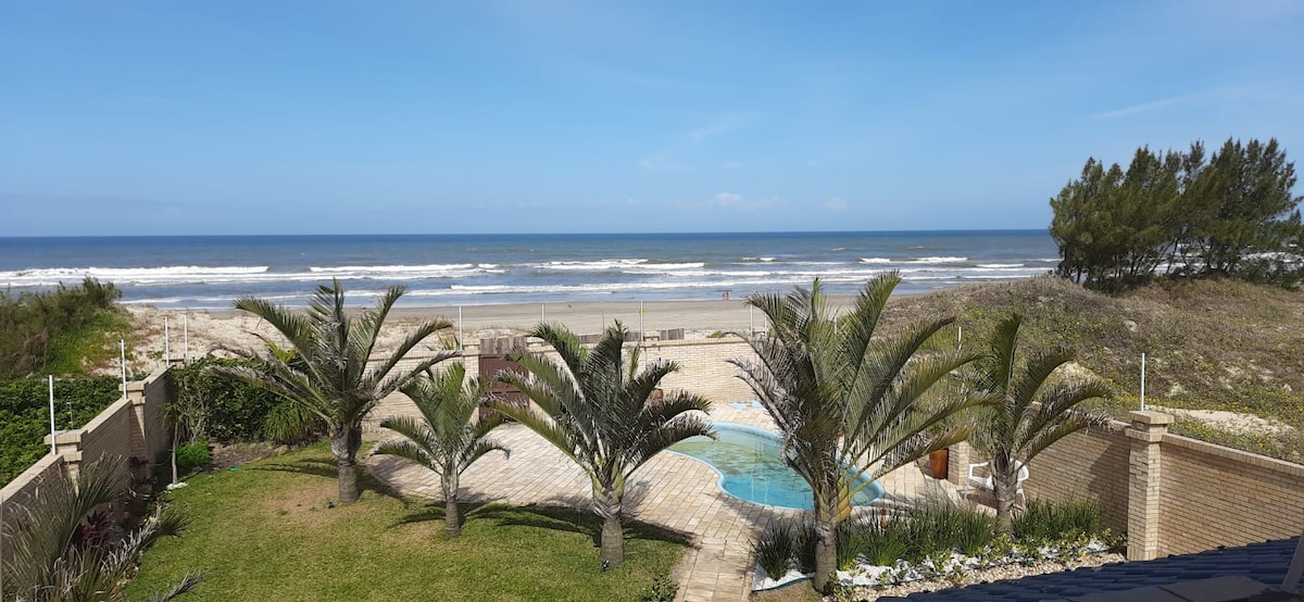 A scenic view captures a tranquil beach bordered by lush greenery and palm trees. The ocean waves gently lap against the shore, and a clear blue sky spans above. A pool area is visible in the foreground, adding to the serene coastal atmosphere.