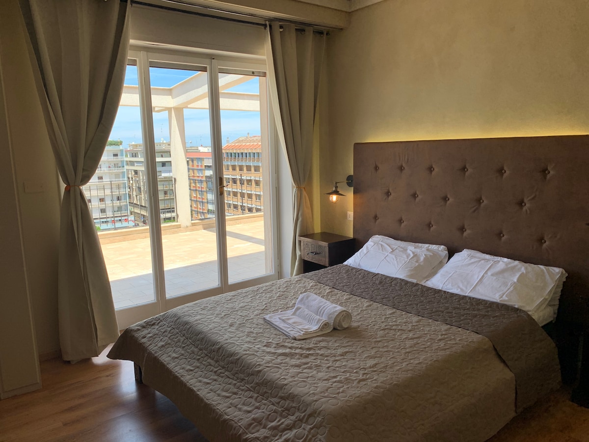 A well-appointed bedroom features a spacious bed with neutral bedding, complemented by a bedside table. Large glass doors provide access to a private terrace, allowing natural light to fill the room and offering views of the surrounding area.