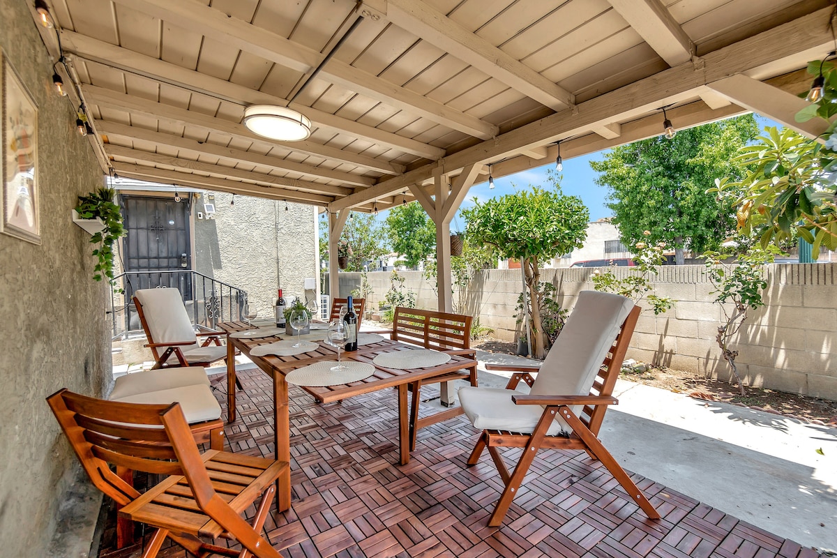 A covered outdoor space features wooden patio furniture, including several lounge chairs and a dining table. The area is bordered by greenery and partial walls, creating a private atmosphere. Natural light filters through the overhead beams, enhancing the inviting setting for relaxation.