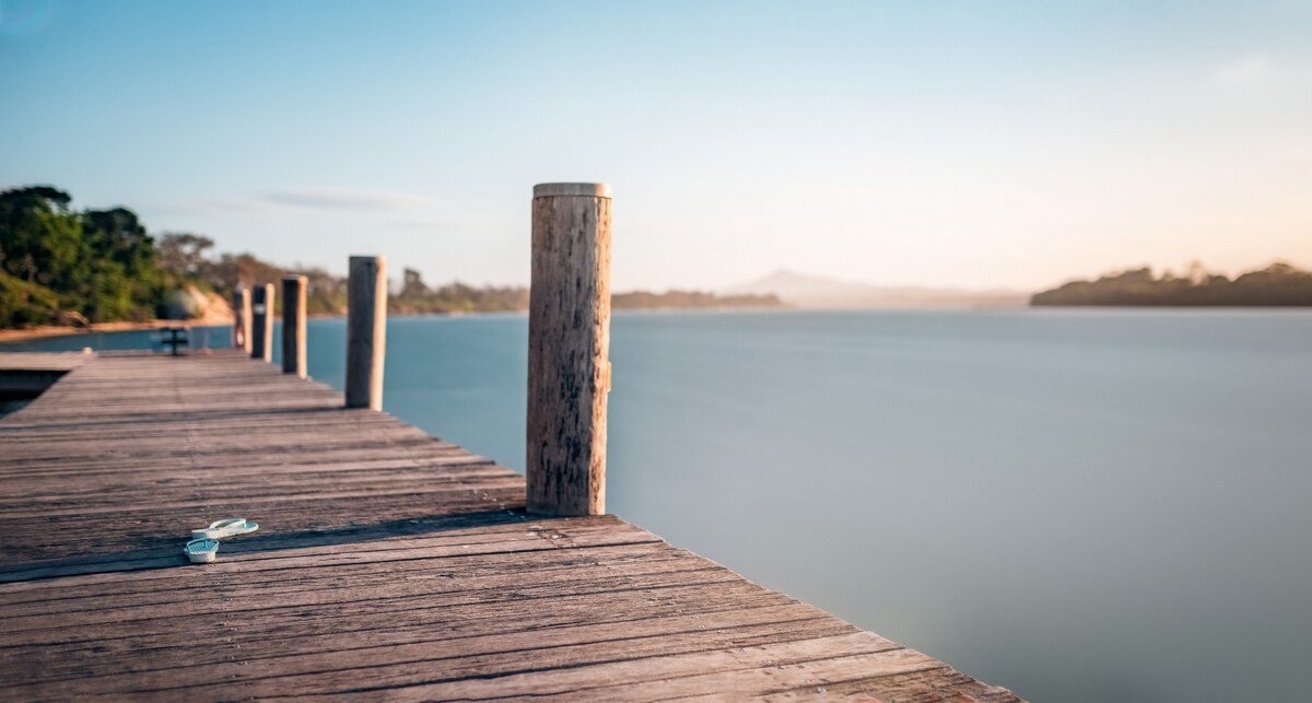 A wooden dock extends over calm waters, supported by several sturdy posts. The serene landscape is enhanced by gentle hills in the background, and the sky displays a soft gradient of colors as daylight transitions. The scene conveys a peaceful atmosphere, ideal for relaxation.