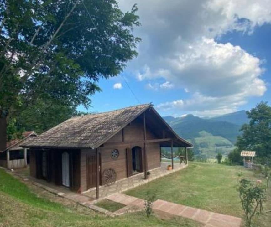 A wooden cabin is nestled on a grassy hillside, surrounded by trees. The structure features a sloped roof and a covered porch. Expansive views of distant mountains and clouds are visible in the background, enhancing the natural setting.