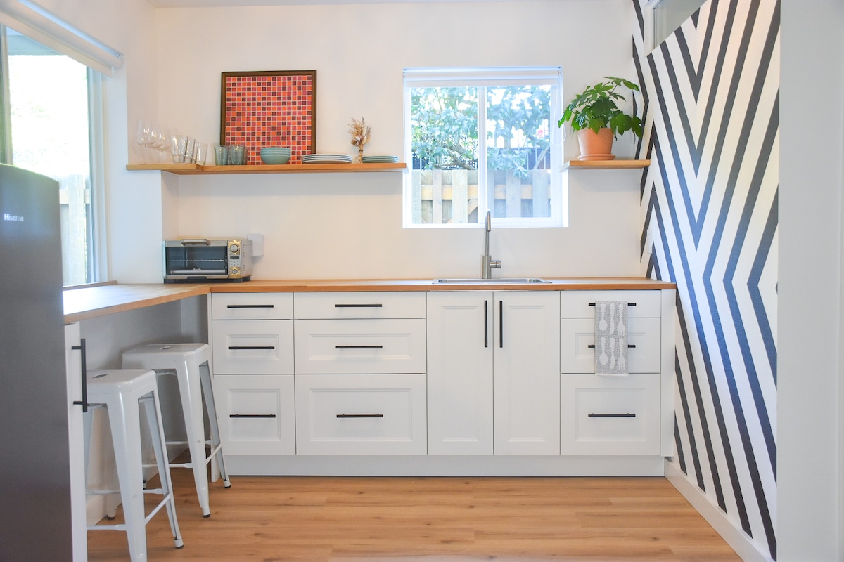 A modern kitchen is presented with sleek white cabinetry and wood countertops. Open shelving displays dishware and a potted plant. Natural light filters through a window, while a striped wall adds a graphic element to the space. Two metal stools are positioned at the bar.