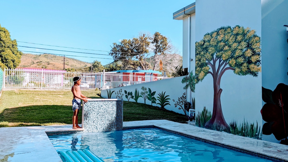 A serene outdoor space features a sparkling pool, surrounded by vibrant greenery and artistic wall murals. A water feature is visible, with a child standing nearby, gazing at the inviting surroundings under a clear blue sky.