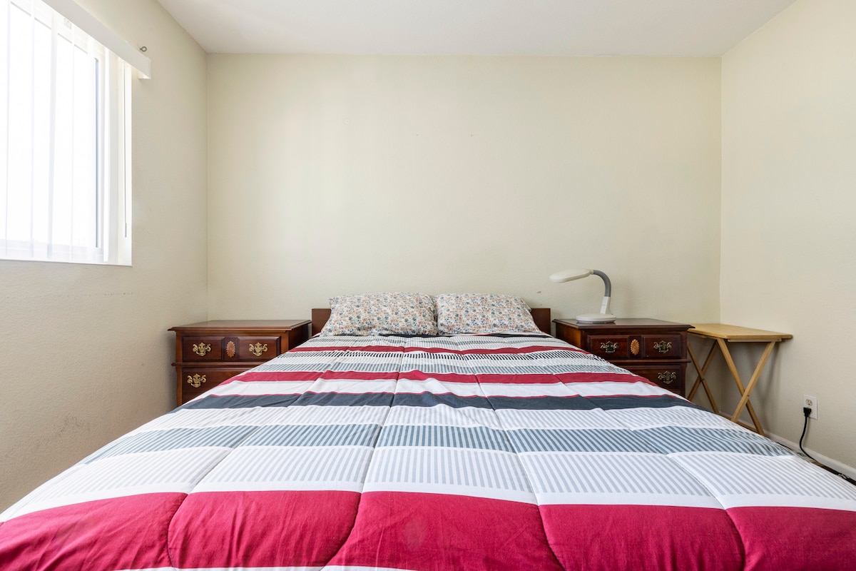A spacious bedroom features a large bed with a patterned bedding set. Two bedside tables flank the bed, and a small folding table is positioned beside a window that allows natural light to enter the room.