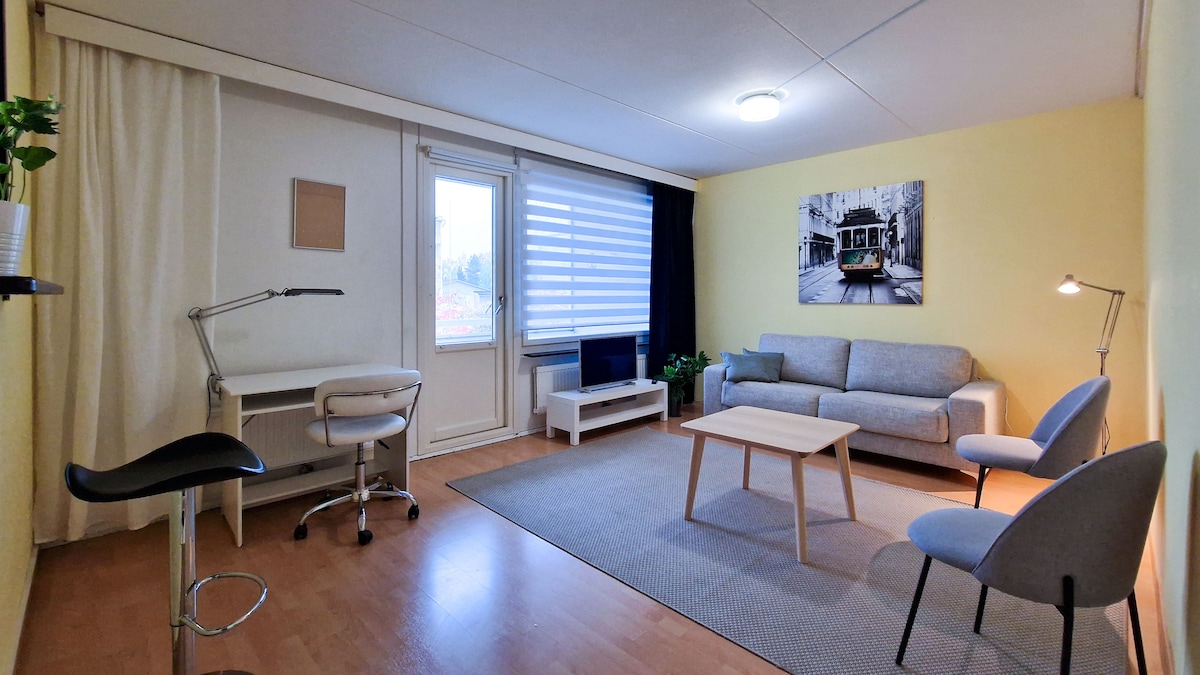 A spacious living area features a comfortable grey sofa, two light blue chairs, and a wooden coffee table centered on a woven rug. A work desk with a chair is situated against a wall. Natural light enters through large windows, adorned with partially closed white blinds.