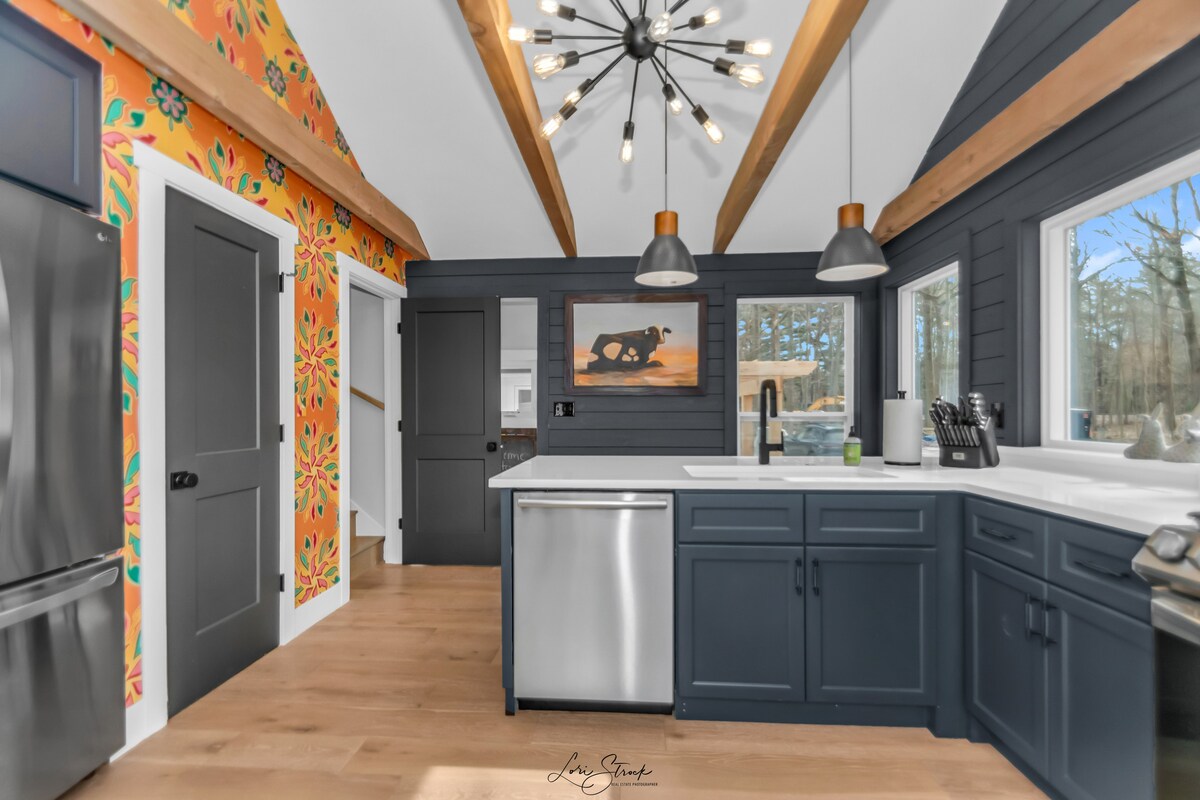 The kitchen features modern appliances, including a stainless steel refrigerator and dishwasher. Dark cabinetry contrasts with a vibrant floral accent wall. Exposed wooden beams enhance the ceiling, and large windows offer views of the surrounding forest.