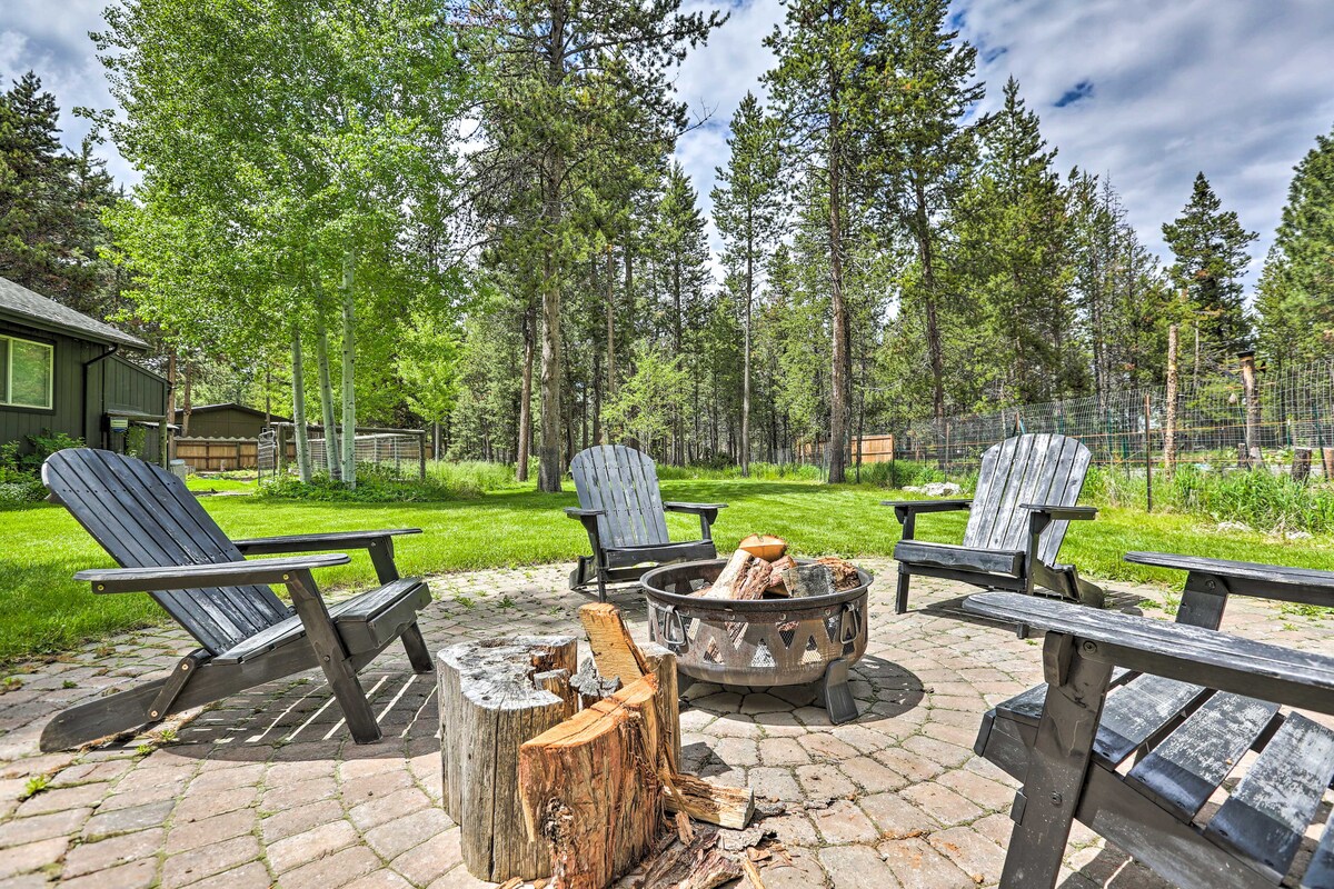 A spacious outdoor area is surrounded by lush green trees, featuring a circular fire pit with natural stone edges. Four Adirondack chairs are positioned around the fire pit, offering a cozy place to gather. A grassy yard extends beyond, providing ample space for relaxation.