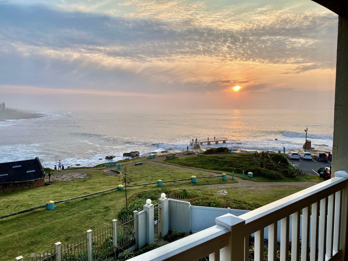 A serene early morning view captures the sun rising over the ocean, illuminating the misty landscape. The horizon features hues of orange and soft pink, while the foreground displays a grassy area leading to the beach. A small wooden pier extends into the water, enhancing the coastal scene.