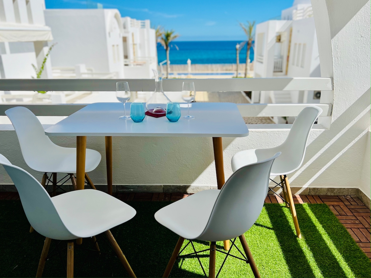 A dining table with a white surface and wooden legs is set with glasses and a carafe, overlooking the Mediterranean Sea. Four white chairs surround the table, positioned on a green grass-like surface, with palm trees visible in the background.