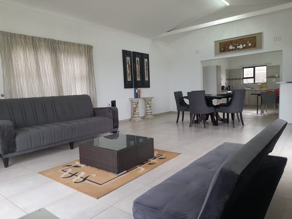 A spacious living area is showcased with gray sofas positioned around a low coffee table. A decorative area rug lies beneath the table. Natural light floods the room through large windows covered by sheer curtains, enhancing the bright ambiance of the open space.