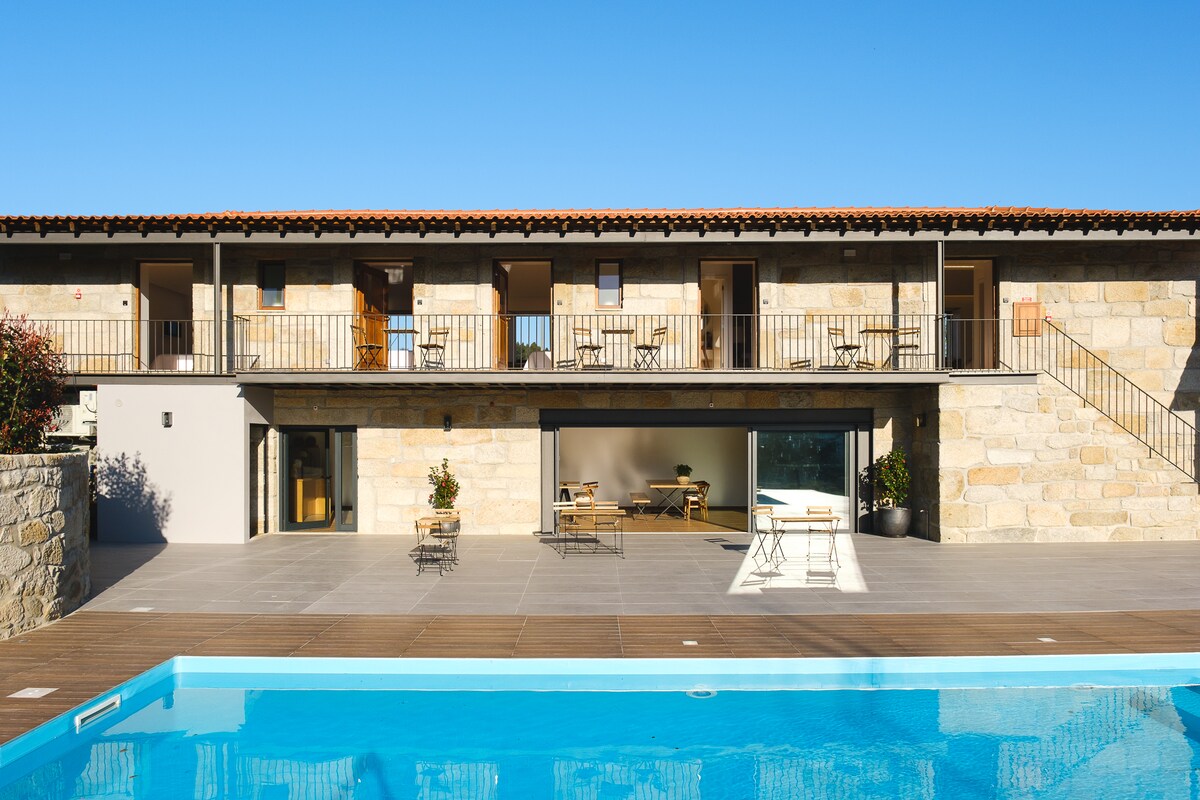 A stone building features a balcony with several chairs, overlooking a clear blue pool. The open glass doors reveal a bright indoor space, while the patio area is designed with minimalist furniture. The landscape is complemented by a clear blue sky.