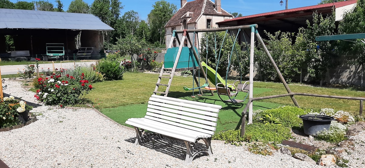 A well-maintained garden area features a white bench amidst colorful flower beds. A swing set with two swings is present, along with a grassy area for relaxation. The surrounding greenery creates a peaceful outdoor space, enhancing the overall ambiance of the property.