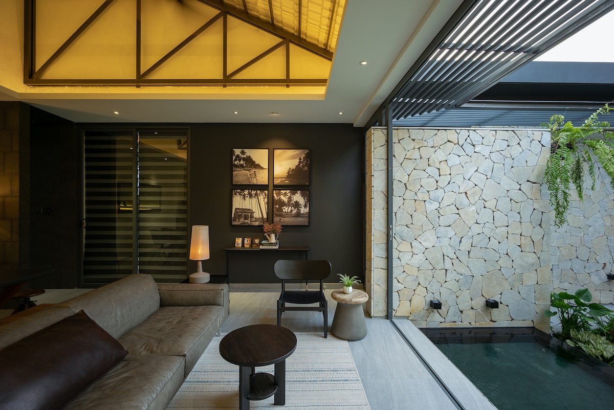 A modern living area features a comfortable sofa, a round wooden coffee table, and a potted plant. Large windows allow natural light to illuminate the space, with stone walls adding texture. A serene pool is visible, enhancing the indoor-outdoor connection.