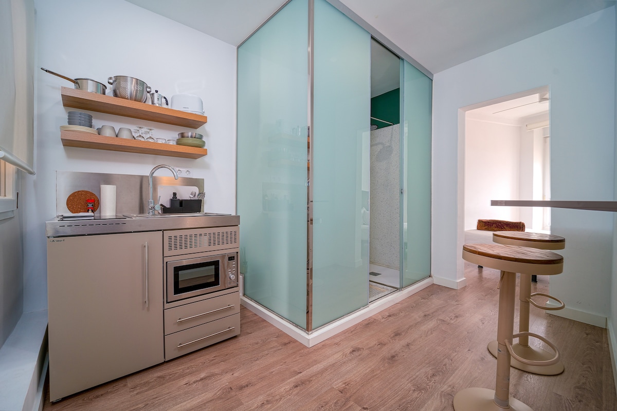 A compact kitchen area features a microwave, stove, and sink, positioned beside a glass-enclosed shower. Overhead shelves hold dishes and utensils, while a spacious counter with high stools offers a dining or workspace. Natural light enters through a nearby window, brightening the modern space.