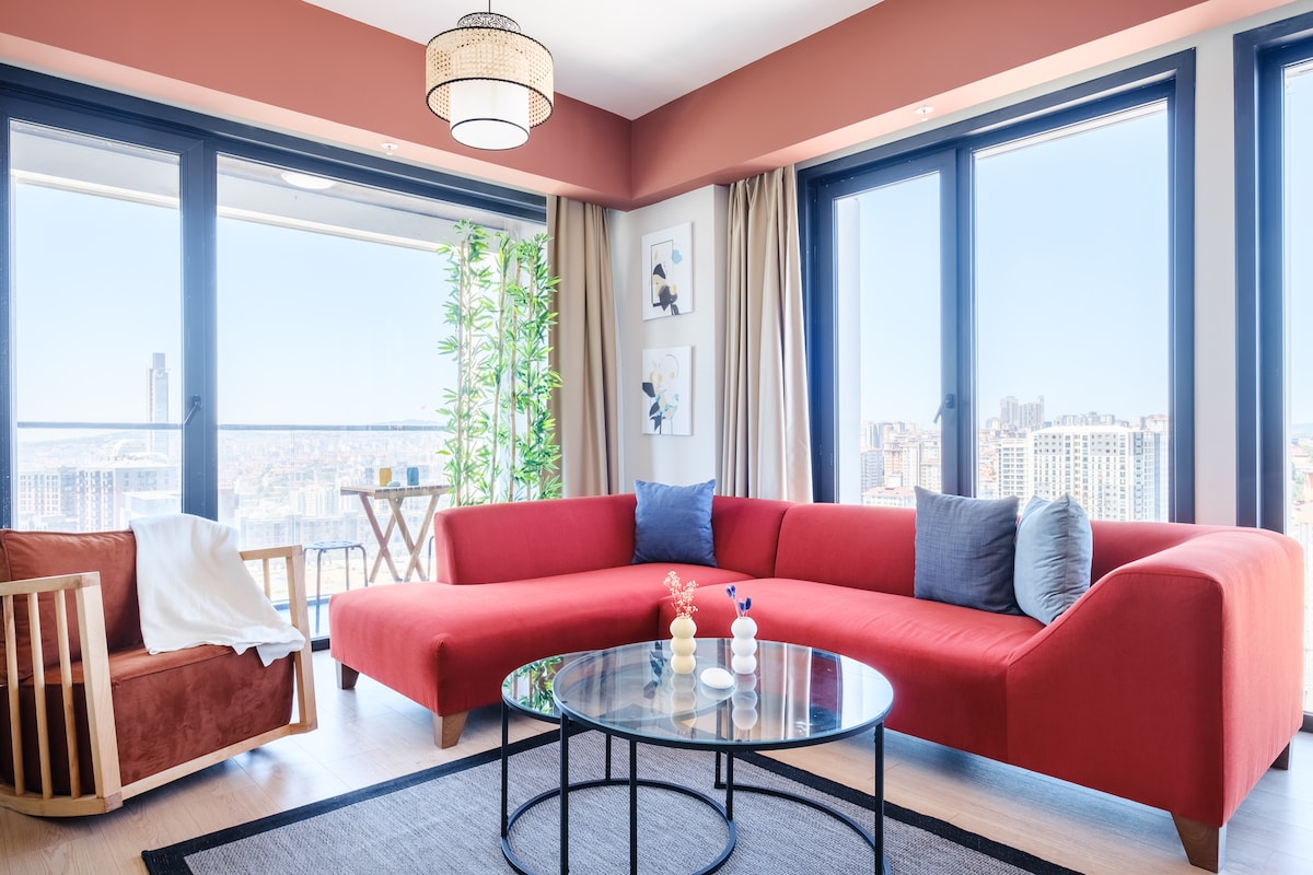 A modern living area features a sectional red sofa and a wooden chair, offering ample seating with a city view through large glass windows. A round glass coffee table is positioned in the center, complemented by minimalistic decor on the walls.