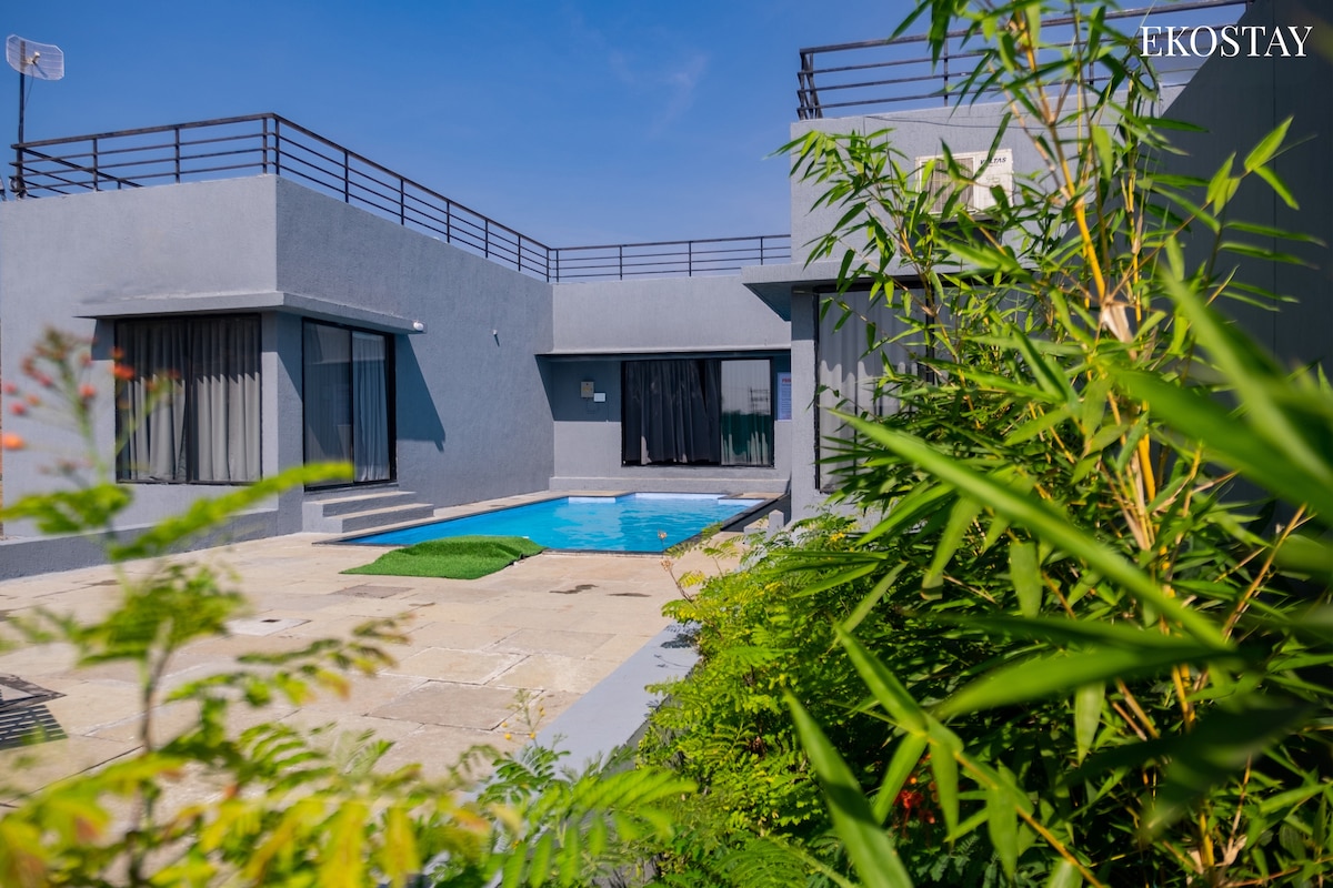 A private pool is visible against a backdrop of modern villa architecture, framed by lush greenery. Sliding glass doors open to a spacious patio area, providing a seamless connection between indoor and outdoor spaces. The sunlit atmosphere enhances the inviting appeal of this secluded retreat.