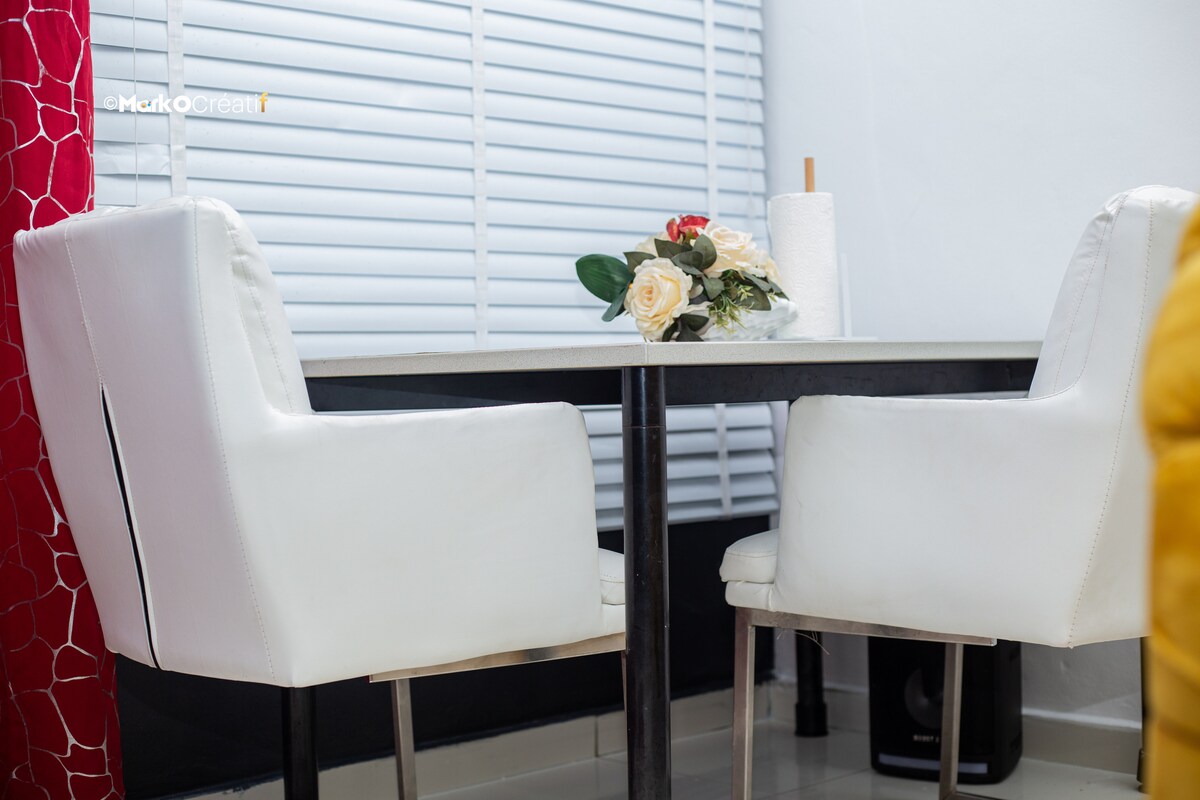 A dining area is presented with a sleek, modern table and two white upholstered chairs. A small vase with roses is positioned centrally on the table. Light filters through partially closed window blinds, adding brightness to the space.