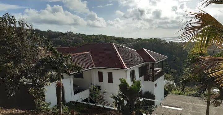 A spacious three-bedroom house is situated on a hillside, featuring a terracotta roof. Surrounding greenery and tropical plants enhance the serene environment, with panoramic views of the landscape visible from the property. The ocean can be seen in the distance beneath a partly cloudy sky.