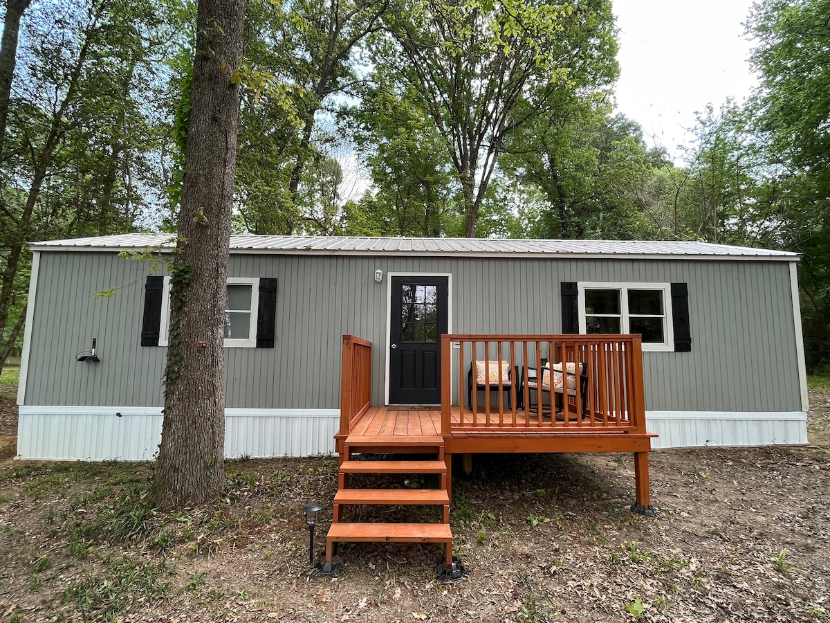 The exterior of a modern cabin is highlighted, featuring a wooden deck with seating arrangements. Large windows are framed by black shutters, and a welcoming dark front door is centrally positioned. Surrounding greenery adds to the natural setting.