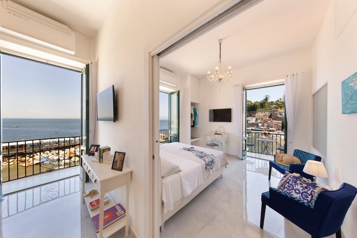 A light-filled bedroom is showcased, featuring a spacious bed adorned with fresh white linens. Two large windows frame views of the sea and the fishing village below, while a cozy armchair and a small side table are positioned nearby, enhancing the room's comfort.
