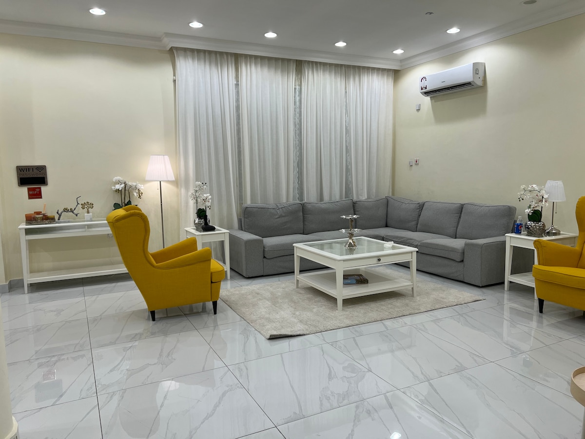 A bright living area features a large gray sectional sofa complemented by two yellow armchairs. A coffee table sits on a soft rug in the center, surrounded by light-colored walls and elegant curtains. Natural light enhances the modern decor.