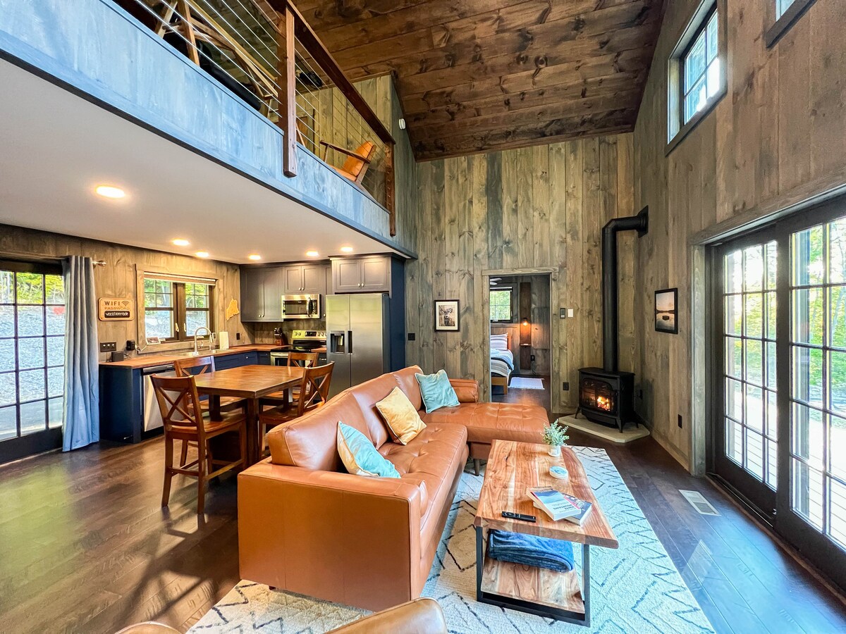 The living area features a spacious leather sofa, complemented by accent pillows. A wooden coffee table occupies the center, while a Vermont Casting gas stove adds warmth. Large windows allow natural light to fill the room, with the kitchen and dining area visible in the background.