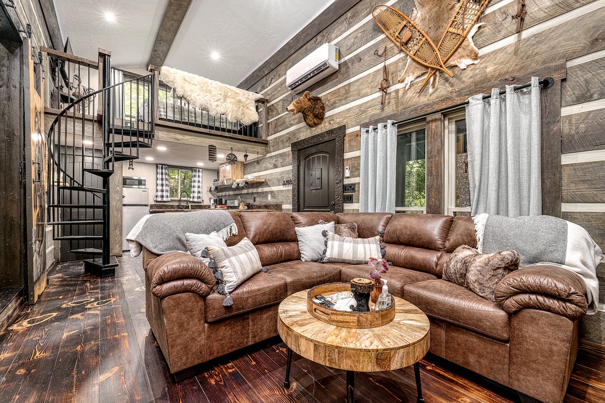 A spacious living area features a large brown sectional sofa adorned with cozy pillows and throws. A circular wooden coffee table is positioned in front, complemented by decorative accents. Natural light filters through sheer curtains, highlighting the rustic textures of the wall and staircase.