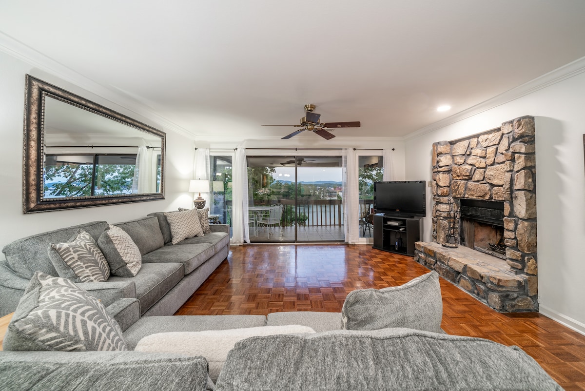 A spacious living area is presented with a large sectional sofa arranged around a coffee table. A stone fireplace stands as a focal point, and a wall-mounted television is positioned nearby. Glass doors open to a view of Lake Hamilton, enhancing the area’s natural light.