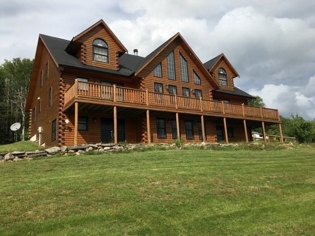 A large log home features a spacious front deck, surrounded by grassy areas and rocky landscaping. The structure boasts multiple gabled roofs and large windows, allowing natural light to fill the interior. A satellite dish is mounted on one side.