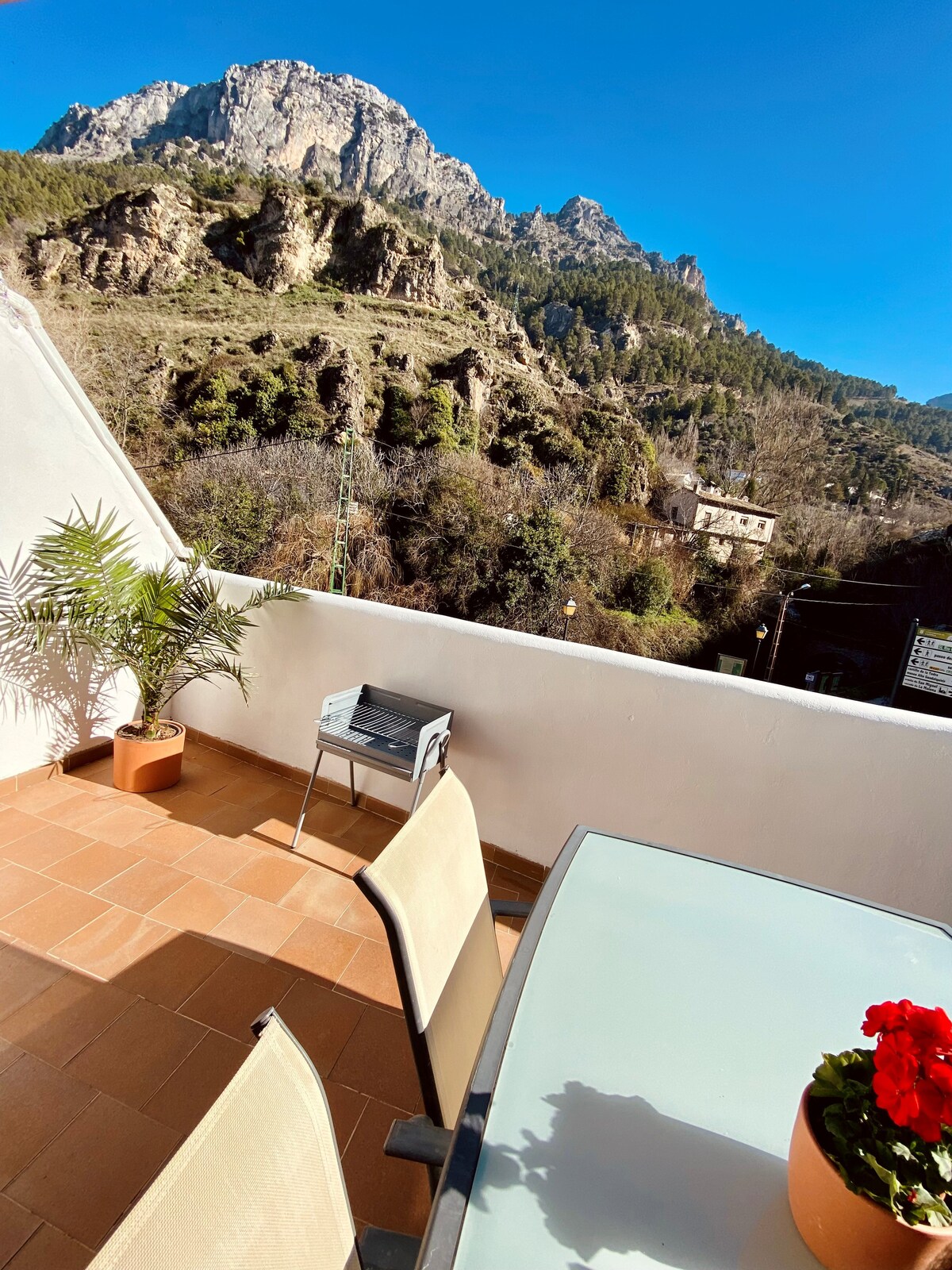 An outdoor terrace is featured, furnished with a glass table and two chairs, overlooking a mountainous landscape. A potted plant adds a touch of greenery. The clear blue sky enhances the vibrant setting, creating a peaceful area for relaxation.