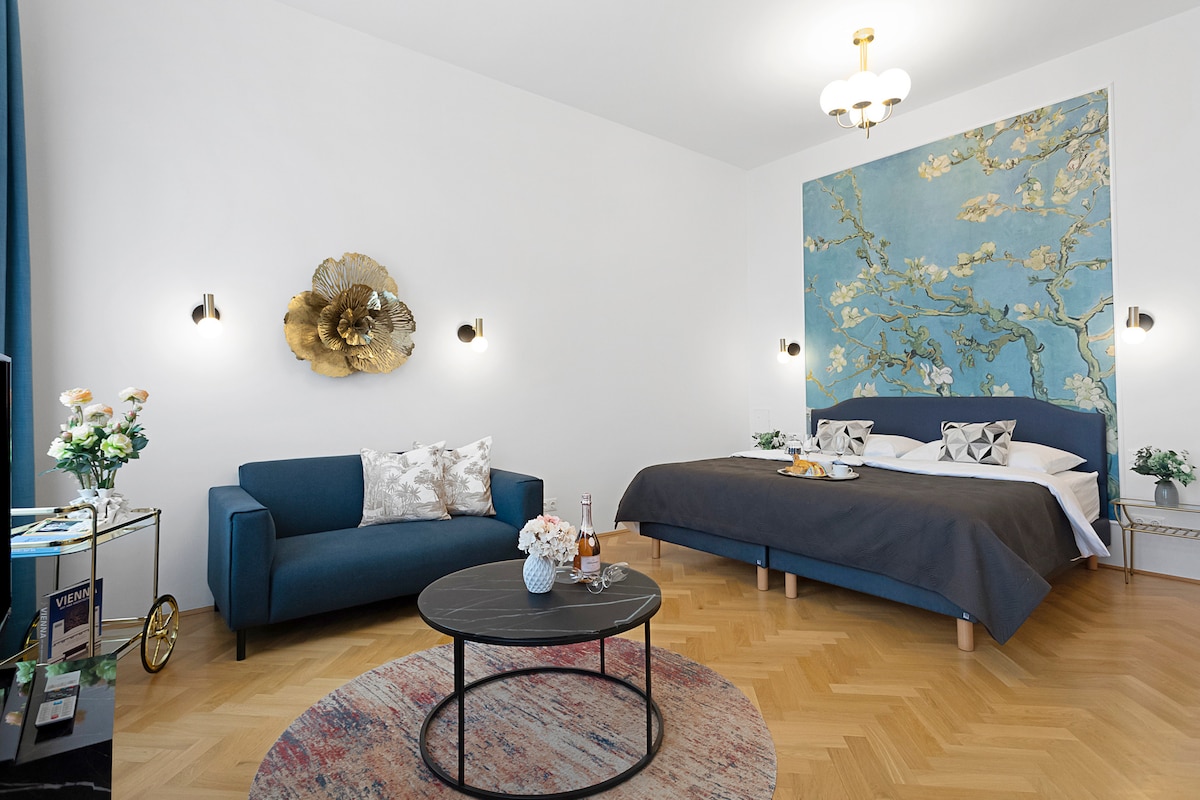 A spacious room features a large kingsize bed with elegant bedding and decorative pillows. A cozy blue sofa is positioned next to a round coffee table, accented by floral arrangements. An artistic blue wall hanging adds character, while soft lighting is provided by wall sconces.