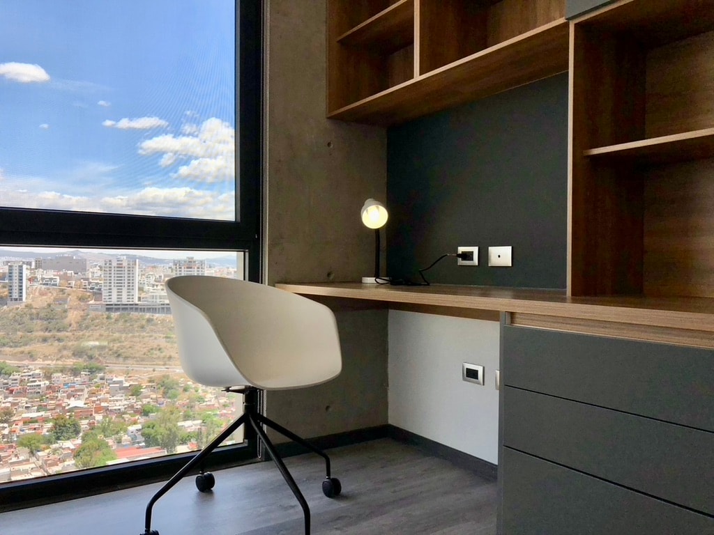 A well-lit workspace features a modern desk with a stylish chair. Natural light floods the area through the large window, revealing a view of the city below. Minimalistic shelves are positioned above the desk for storage.