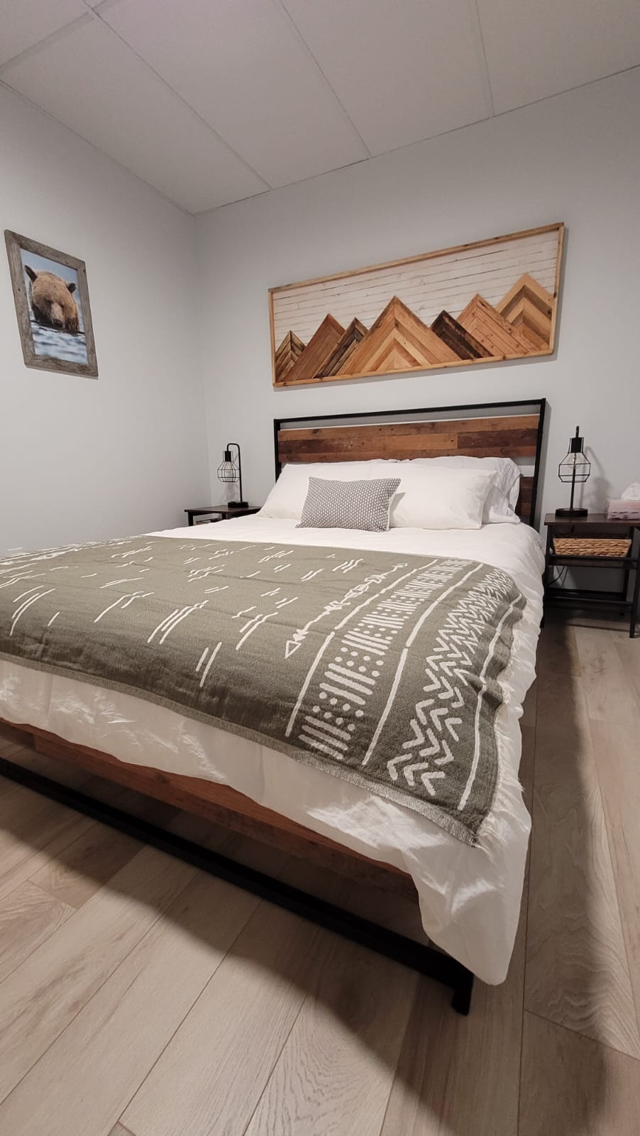 A serene bedroom features a spacious bed with a textured blanket draped across. Light wood flooring complements the room's neutral tones. Framed mountain art and a wildlife picture adorn the walls, adding character, while bedside tables with lamps provide a cozy touch.