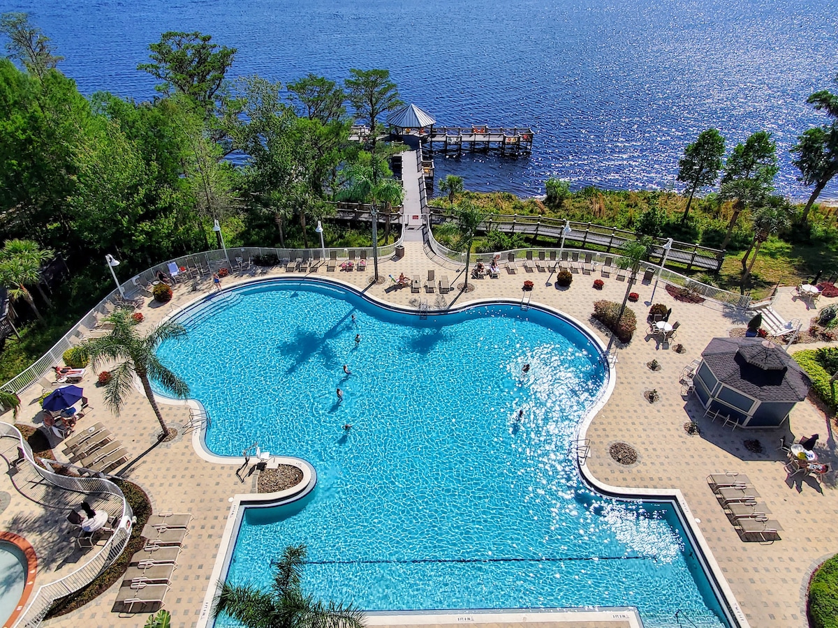 An expansive swimming pool lies beneath clear blue skies, featuring a unique shape surrounded by lounge chairs and umbrellas. Lush greenery frames the pool area, and a boat dock extends into the shimmering waters of Lake Bryan, offering picturesque views.