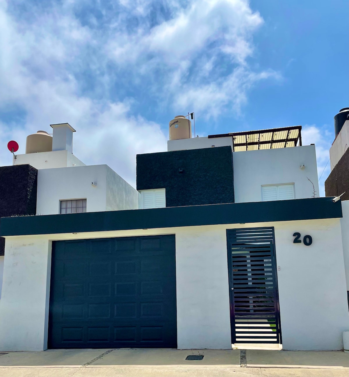 The exterior of the house features a modern design with a combination of light and dark walls. A blue garage door is visible, along with a decorative metal screen. A rooftop terrace is situated above, and a clear blue sky adds to the overall setting.