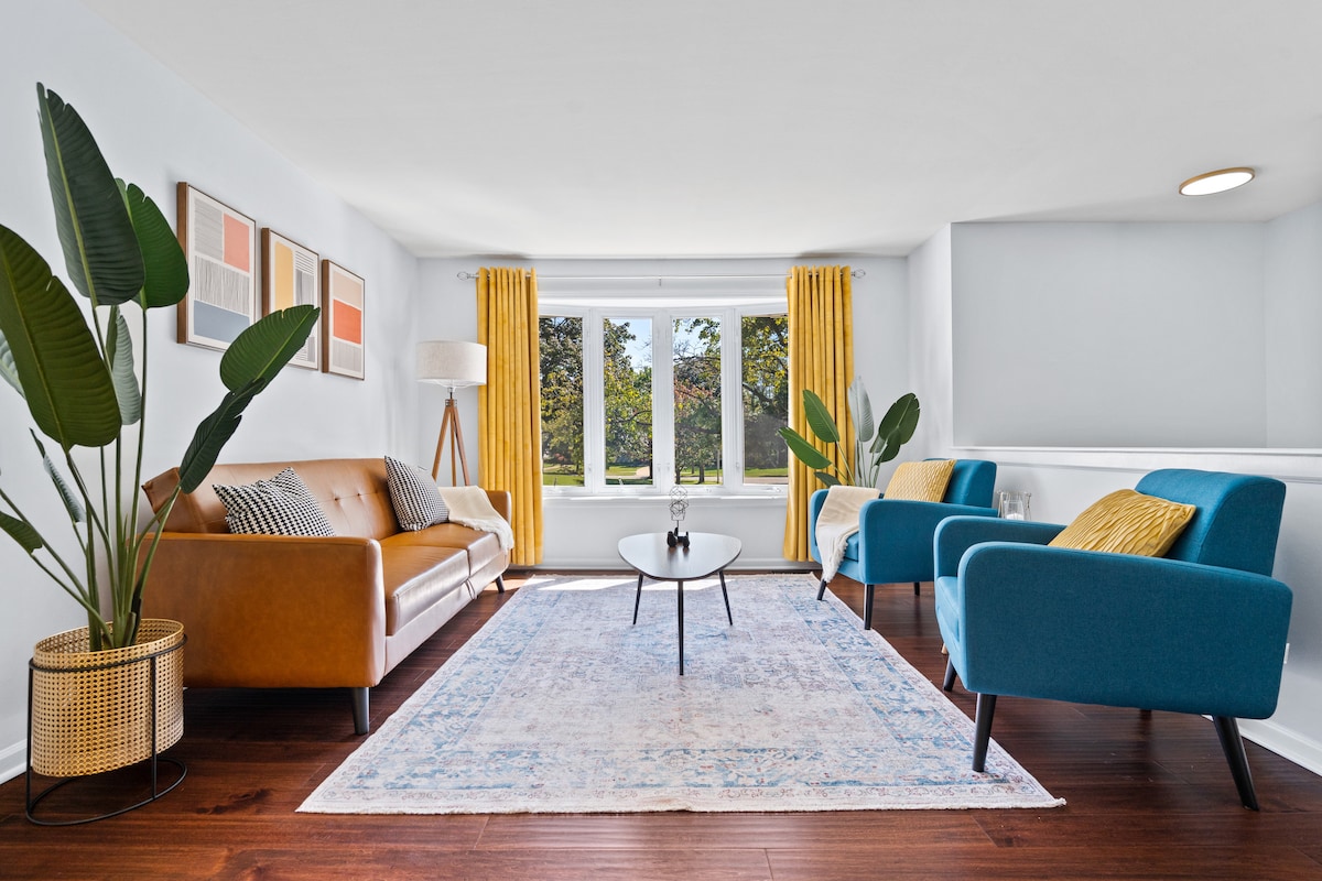 A bright living area features large windows draped with yellow curtains, allowing natural light to fill the space. A tan leather sofa and two blue chairs are arranged around a round coffee table, with a light area rug underfoot and decorative wall art adding a modern touch.