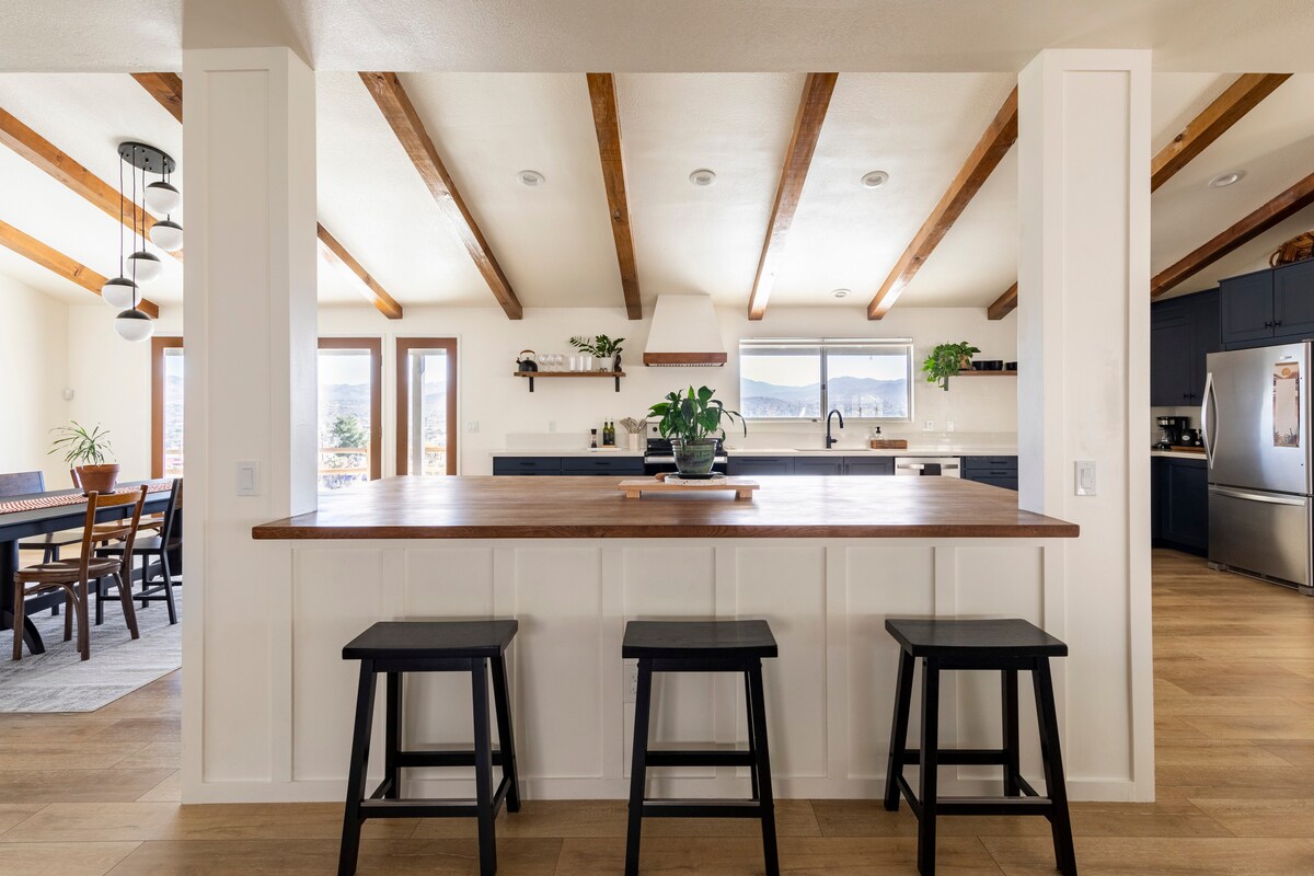 An open kitchen and dining area features exposed wooden beams and a center island with three black stools. Natural light floods the space through large windows, offering views of the surrounding landscape. The kitchen is equipped with modern appliances and ample counter space.