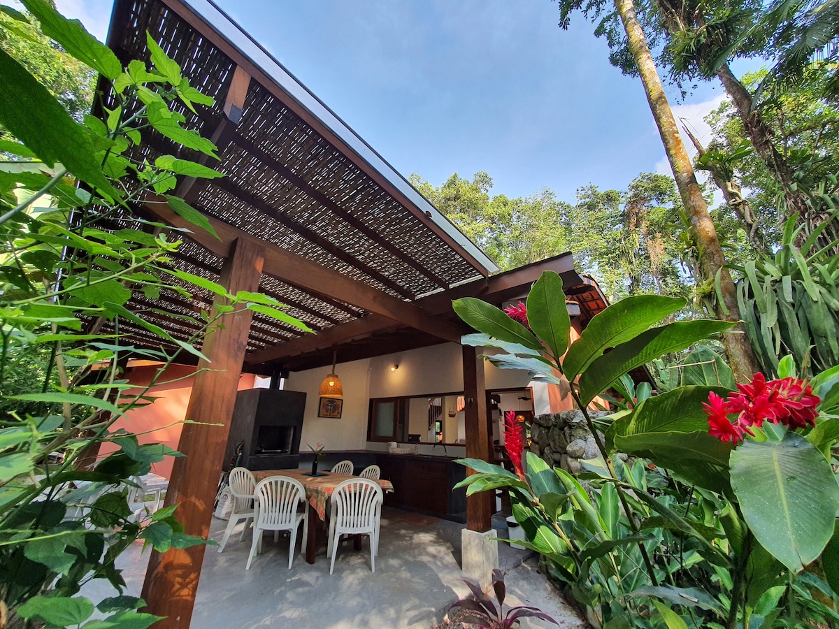 An outdoor seating area is surrounded by lush greenery, featuring a shaded patio with several white chairs and a wooden table. The open design allows for natural light to flow in, with a glimpse of the interior living space visible through the entrance.