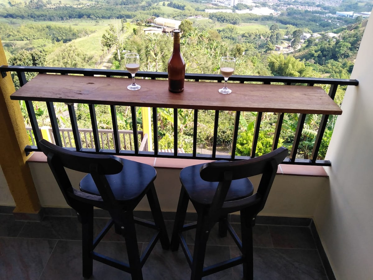 A balcony is showcased with two black high stools positioned next to a wooden bar table. Two glasses are placed on the table, with a bottle of beverage in the center. Lush greenery and hills are visible in the background, contributing to a serene view.