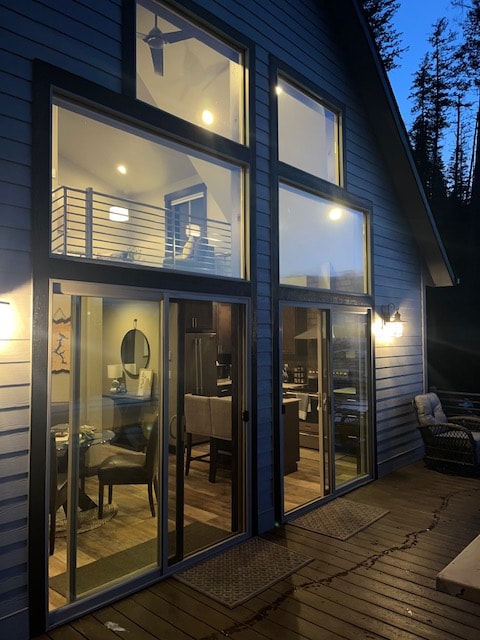 A modern two-story cabin is viewed from the deck, featuring large glass doors that brighten the interior. Warm lighting illuminates the living area and dining space, with a staircase leading to an upper level. Surrounding trees frame the picturesque setting as evening settles in.