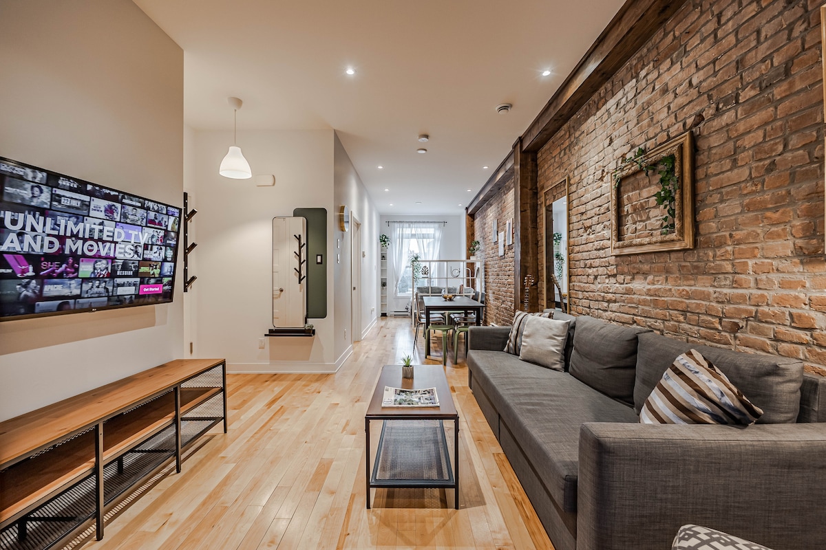A spacious living area features exposed brick walls complemented by hardwood floors. A comfortable sofa is placed in front of a mounted flat-screen TV, and a coffee table is positioned nearby. Natural light enters through a window, illuminating the inviting space.