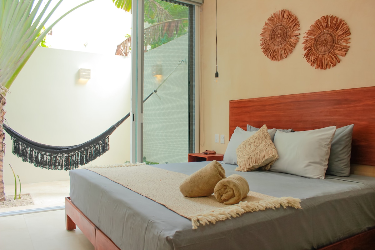 A spacious bedroom features a large bed adorned with neutral-toned linens. Decorative wall hangings are displayed above the bed. Natural light filters in through the glass doors, leading to an outdoor area with a hammock and lush greenery.