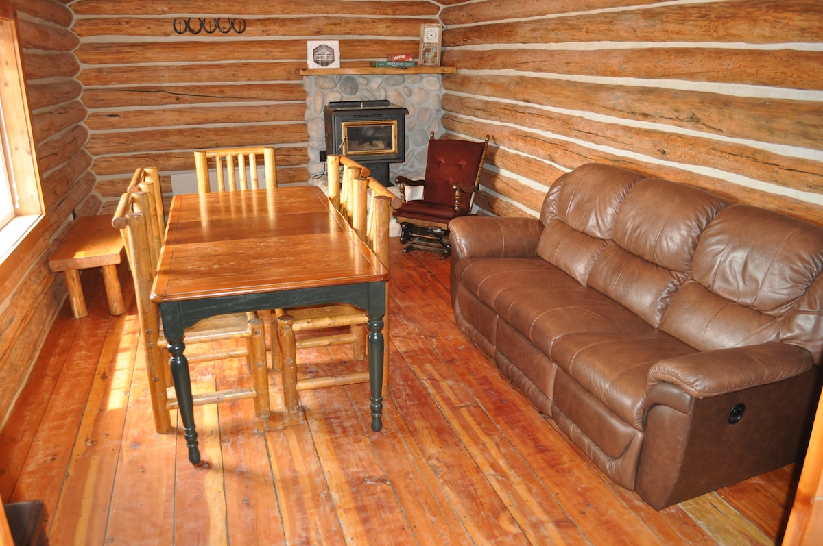 A rustic interior space features a wooden dining table surrounded by five chairs, complemented by a brown leather couch. A stone fireplace serves as a focal point, while natural light enters through a window. The wooden floor adds warmth to the cabin atmosphere.